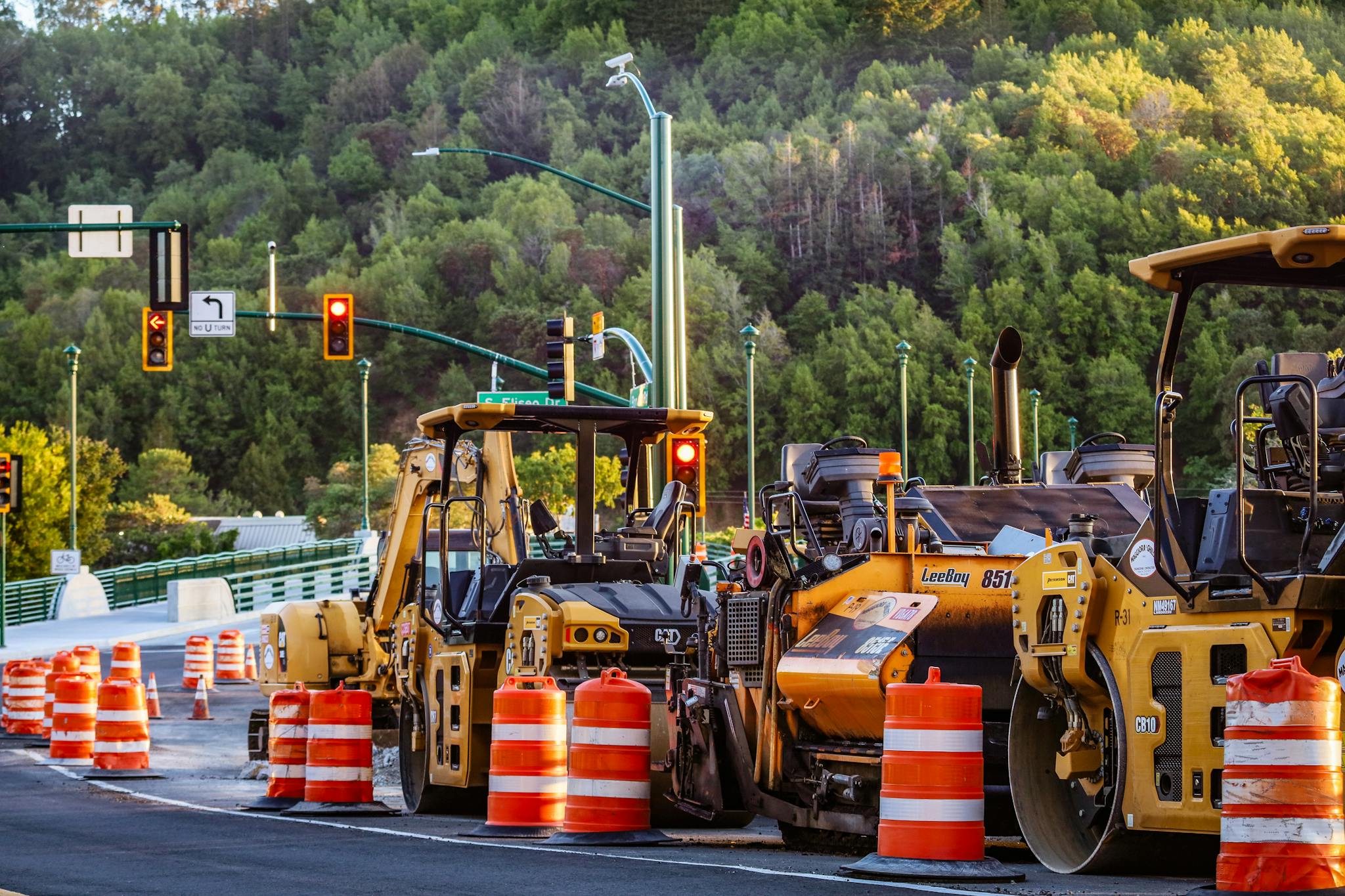 Heavy machinery and barriers at a road construction site with traffic lights and forest backdrop.