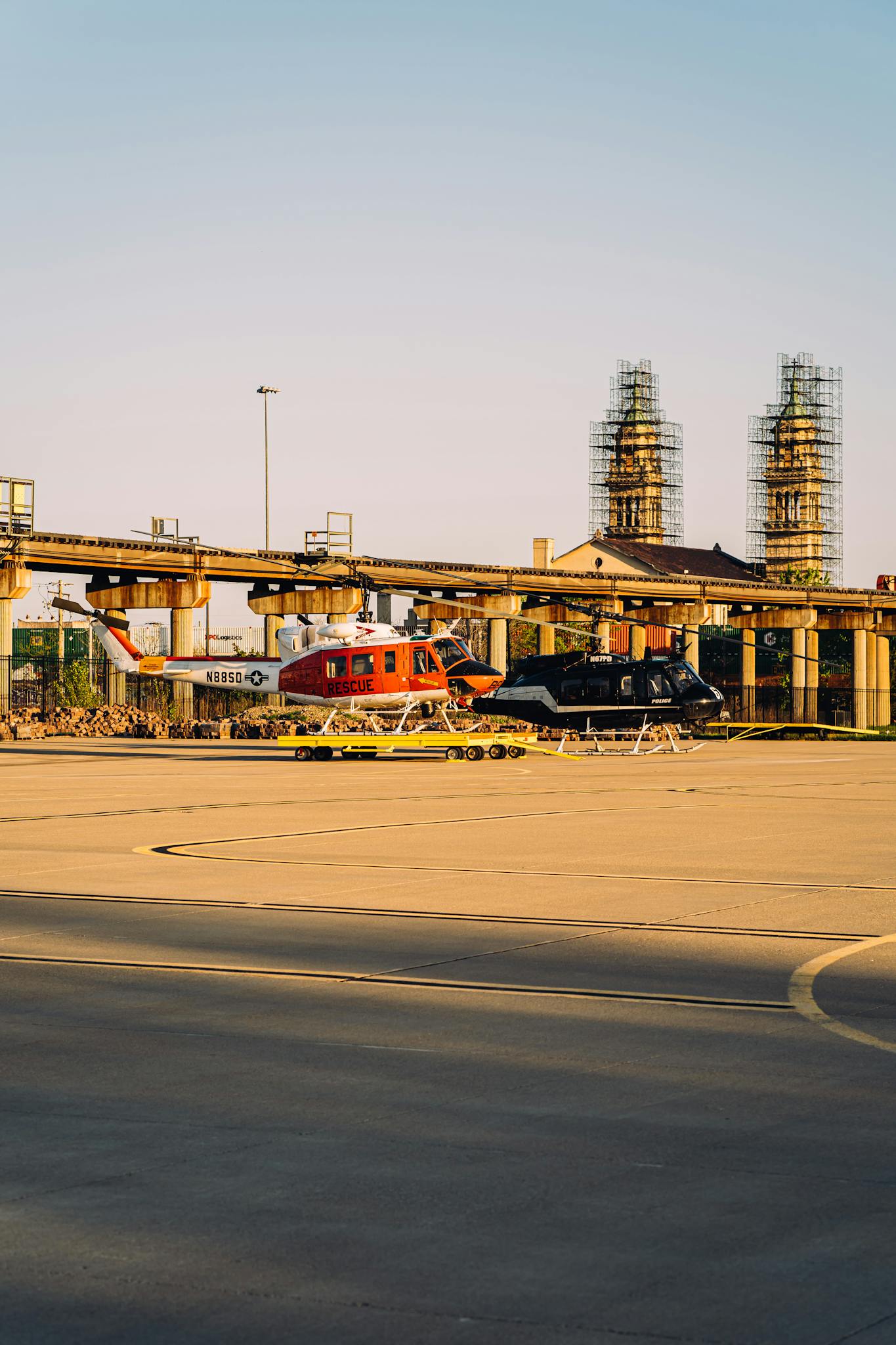 Two helicopters parked on a helipad in an urban industrial area during sunset.